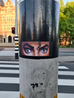 Close-up of a sticker featuring Michael Jackson's eyes. The sticker is affixed to a pole on a city street.
