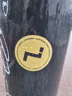 Street sticker Round yellow sticker with a stylized 'L' logo in black. The text around the logo reads:  'COMMUNITY OF LESVOS - SYNTHESIS - ELECTRONIC MUSIC COMMUNITY'. The sticker appears to be affixed to a dark-colored pole outdoors and has water droplets on it.