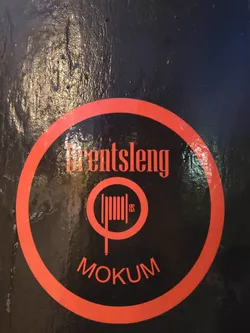Street sticker A round sticker with a red border on a black background. It has the text "Drentslang" at the top, a symbol resembling a coil of rope in the center, and "Mokum" at the bottom. The symbol and text are in red.