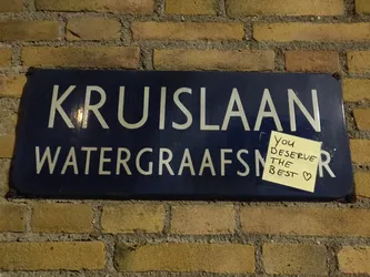 Street sticker A yellow sticky note with the handwritten message "You deserve the best" stuck to a vintage blue street sign. The street sign reads "Kruislaan Watergraafsmeer" in white lettering. The photo was likely taken at night or in low light conditions.