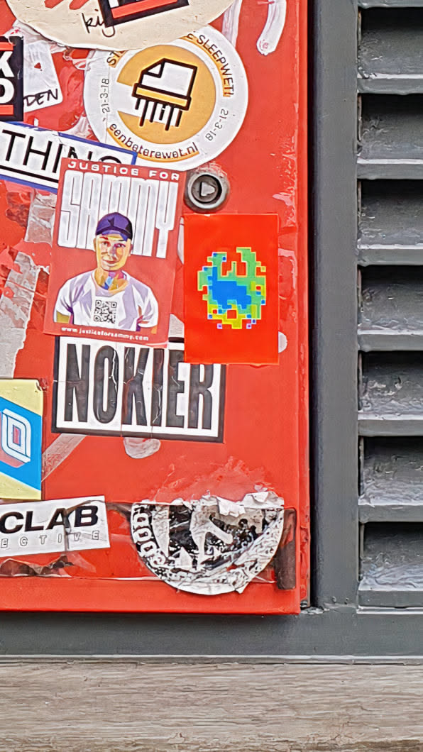 A square red sticker with a pixelated globe in shades of blue, green, and yellow. The design resembles an 8-bit graphic. It is affixed to a red surface alongside other stickers.