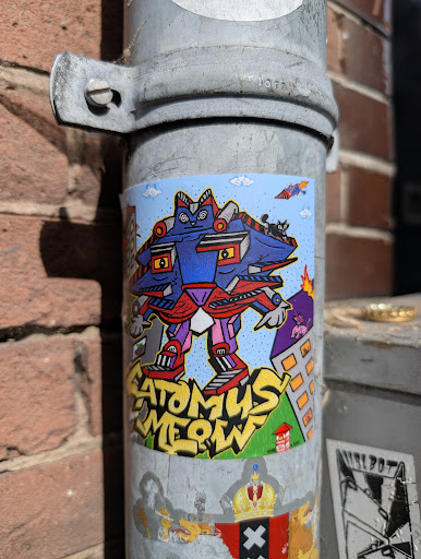 The sticker showcases a whimsical robot with cat-like features, standing tall over a stylized cityscape with cartoonish details like a volcano. The text "ATOMUS MEOW" is present. The style is cartoonish and colorful.