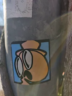 A vinyl sticker featuring a cartoon character with large glasses and a hat. The character has a round, orange head and a simple, charming design. The sticker is affixed to a grey metal pole outdoors.
