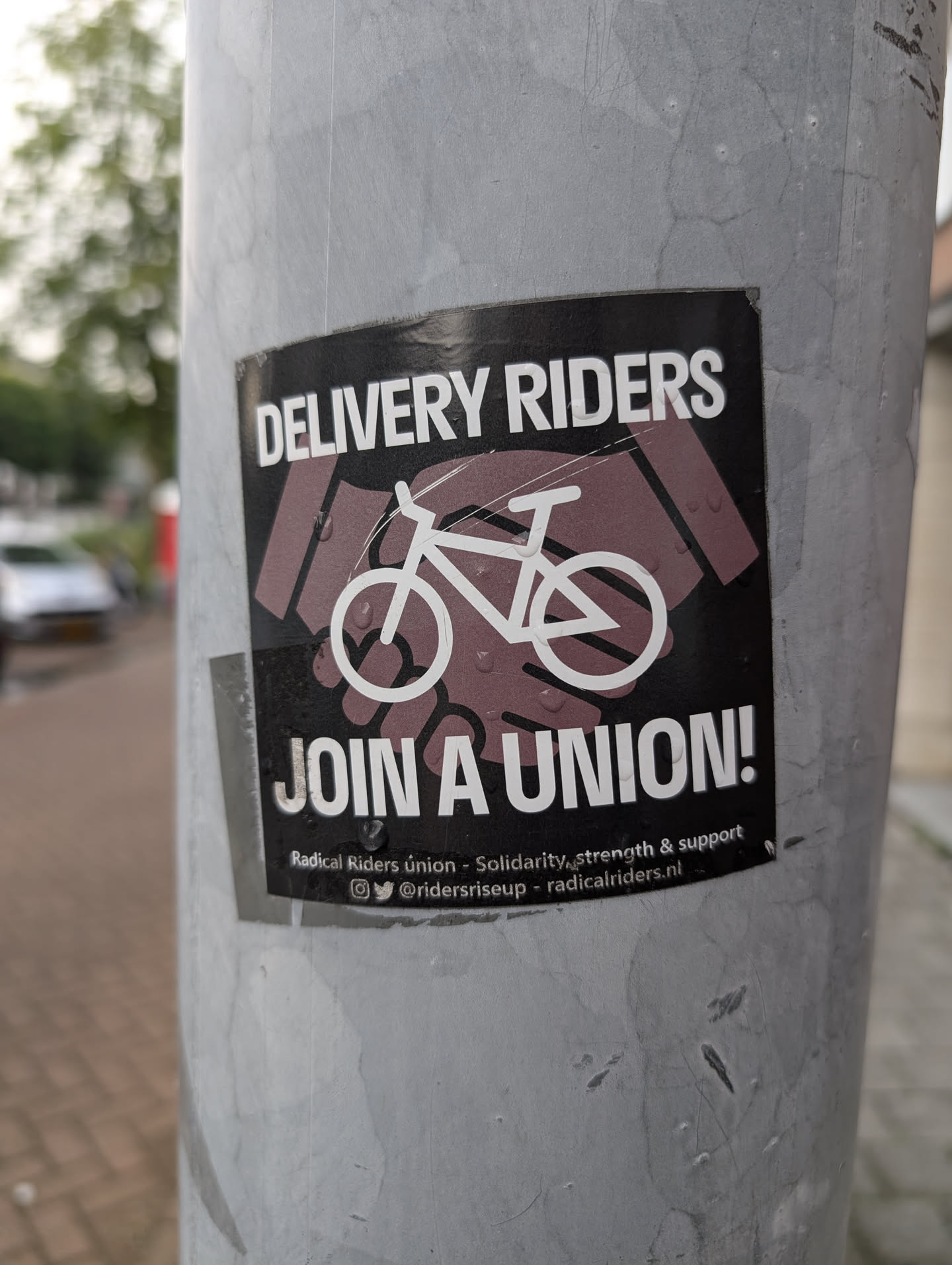 The sticker is black with white text and graphics. At the top, in large white capital letters, it reads 'DELIVERY RIDERS'. Below this, there is an illustration of a handshake with a bicycle silhouette in the center. The handshake is depicted with stylized, dark purple hands. The bicycle is outlined in white. Below the handshake graphic, in large white capital letters, it reads 'JOIN A UNION!'. At the bottom, in smaller white text, it says 'Radical Riders union - Solidarity, strength & support', followed by social media icons and handles. The sticker appears slightly worn and has small water droplets on its surface.
