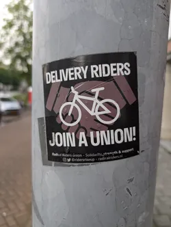 Street sticker The sticker is black with white text and graphics. At the top, in large white capital letters, it reads 'DELIVERY RIDERS'. Below this, there is an illustration of a handshake with a bicycle silhouette in the center. The handshake is depicted with stylized, dark purple hands. The bicycle is outlined in white. Below the handshake graphic, in large white capital letters, it reads 'JOIN A UNION!'. At the bottom, in smaller white text, it says 'Radical Riders union - Solidarity, strength & support', followed by social media icons and handles. The sticker appears slightly worn and has small water droplets on its surface.