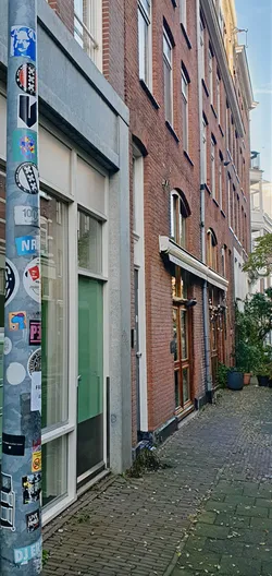 A close-up of a metal pole adorned with various stickers, located on a street in Amsterdam, Netherlands. The stickers feature diverse designs, including the iconic Amsterdam XXX symbol, text, and other graphic elements.