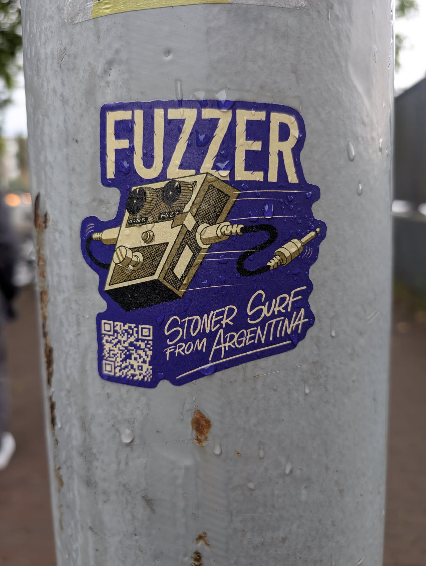 Street sticker A purple sticker with a cut-out shape featuring a vintage-style gold and black guitar pedal. The pedal has two knobs labeled 'TONE' and 'FUZZ', and it's connected by black cables to gold audio jacks. Above the pedal, the word 'FUZZER' is printed in bold, yellow, outlined letters on a blue background. Below the pedal, in smaller yellow text on the blue background, it reads 'STONER SURF FROM ARGENTINA'. A QR code is prominently displayed in the bottom left corner of the sticker.