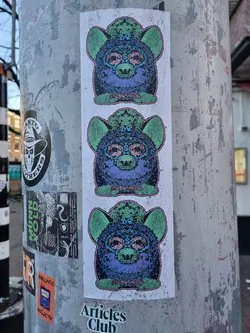 Three identical stickers featuring a glitched, psychedelic Furby. Each Furby has green ears and is wearing a hat with what appears to be a third eye. The text at the bottom of each Furby reads "GELOSHONG". Below the stickers is another sticker that says "Articles Club."