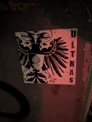 Street sticker The sticker depicts a black and white image of a double-headed heraldic eagle with a shield on its chest containing a pine cone. To the right of the eagle, the word "ULTRAS" is printed vertically in block letters. The sticker is affixed to a textured, possibly concrete, surface.