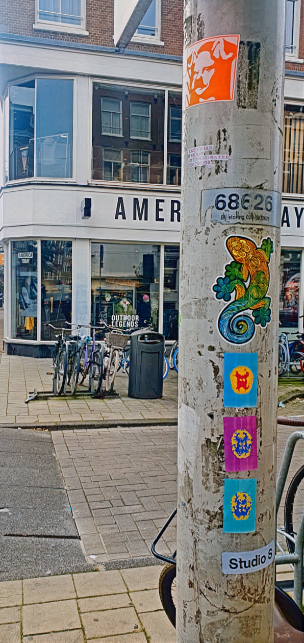 The image shows a metal utility pole covered in various stickers. Centered on the pole is a sticker of a colorful gecko, featuring intricate patterns in shades of orange, yellow, green, and blue. Below the gecko are three square stickers, each with a unique design in bright colors. At the bottom, a sticker says 'Studio S'. The pole is surrounded by a street scene with buildings, bicycles, and a trash bin.