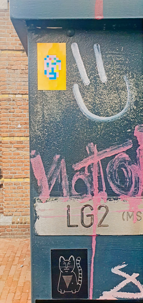A blue metallic post features a yellow square sticker with colorful pixel art. Above it, there is a white painted smiley face. Below, pink graffiti partially covers a plate that reads 'LG2 (MS)'. At the bottom, a black square sticker depicts a white cat.