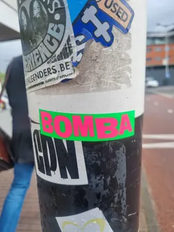 A bright pink and green sticker with the word BOMBA in a bold, uppercase font. It's been placed on a pole that already has several other stickers on it. The pole seems to be located outdoors.