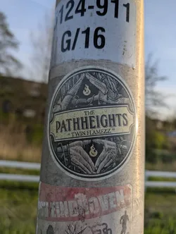 The sticker is circular and features a black and white image of hands forming a pyramid shape at the top and bottom with a flame symbol in the center of each. The text "THE PATH HEIGHTS" is in a rectangular box above the top hand image, and the text "TWIN FLAMEZZ" is below that.
