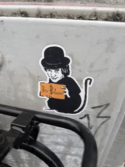 Street sticker The sticker features a black and white illustration of a character wearing a bowler hat and a dark outfit. The character has a distinctive, somewhat unsettling facial expression and is holding up an orange rectangular sign that reads 'Buy Bitcoin' in black text with a stylized dollar sign incorporated into the 'B'. The figure has a tail, resembling a cat or similar animal. The sticker has a white border and appears to be adhered to a textured, light-colored surface.