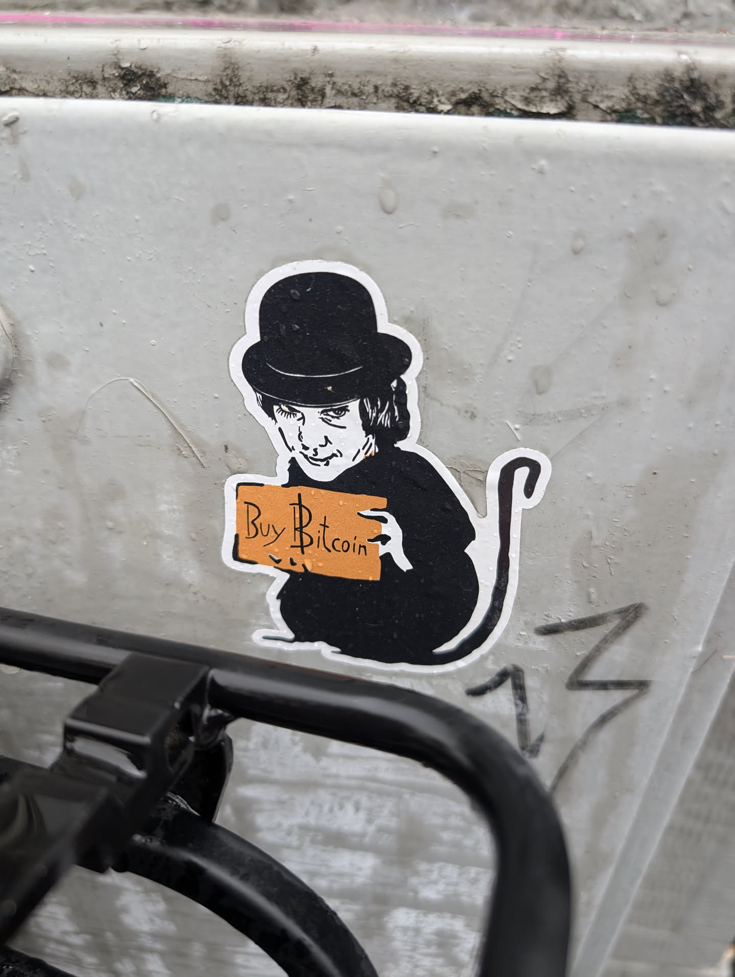 The sticker features a black and white illustration of a character wearing a bowler hat and a dark outfit. The character has a distinctive, somewhat unsettling facial expression and is holding up an orange rectangular sign that reads 'Buy Bitcoin' in black text with a stylized dollar sign incorporated into the 'B'. The figure has a tail, resembling a cat or similar animal. The sticker has a white border and appears to be adhered to a textured, light-colored surface.