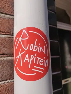 Street sticker A red circular sticker with the words 'Robin Kapitein' written in a stylized white script. The design is simple yet eye-catching. The sticker is affixed to a white cylindrical surface, possibly a pipe or drainpipe.