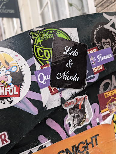 A rectangular black sticker with white cursive text "Lele & Nicola" is adhered to a surface covered in various other stickers. The surrounding stickers have different designs and colors, creating a cluttered and textured background.