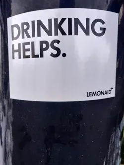 A white sticker on a dark surface with the text 'DRINKING HELPS.' and the logo 'LEMONAID+' at the bottom right corner.
