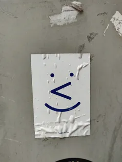 Street sticker A minimalist white sticker features a simple smiley face with a slightly different mouth design.  The sticker is affixed to a textured gray surface. A small 'FRESHCO' logo is visible at the bottom.