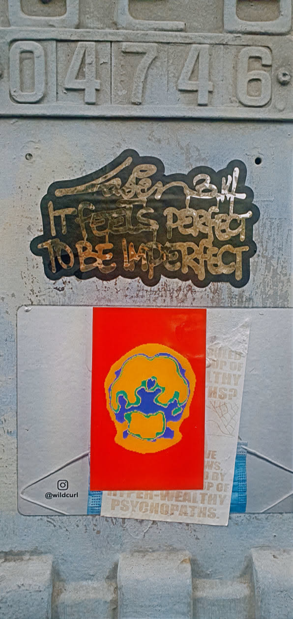The image features a cluster of stickers adhered to a textured, possibly metallic, surface. At the top, the number "04746" is engraved or stamped. The stickers are layered, with one featuring the phrase "It feels perfect to be imperfect". Another sticker has a vibrant red background with an abstract design in yellow, blue, and green. A ripped sticker reveals partial text like "UP OF HEALTHY," "JE NS, BY IP OF," and "UPPER-WEALTHY PSYCHOPATHS."