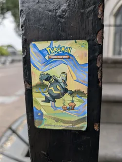 Street sticker The sticker features the Pokémon Gimmighoul in its chest form, crouched low on a grassy, multi-level landscape. Gimmighoul is predominantly dark gray with yellow accents, and it's holding a small treasure chest in its right hand. The background is a vibrant mix of yellow and blue, with streaks of electric-blue energy radiating outwards. The top of the sticker prominently displays the "Pokémon Trading Card Game" logo. The bottom edge of the sticker includes a copyright notice: "©2024 Pokémon / Nintendo / Creatures / GAME FREAK".