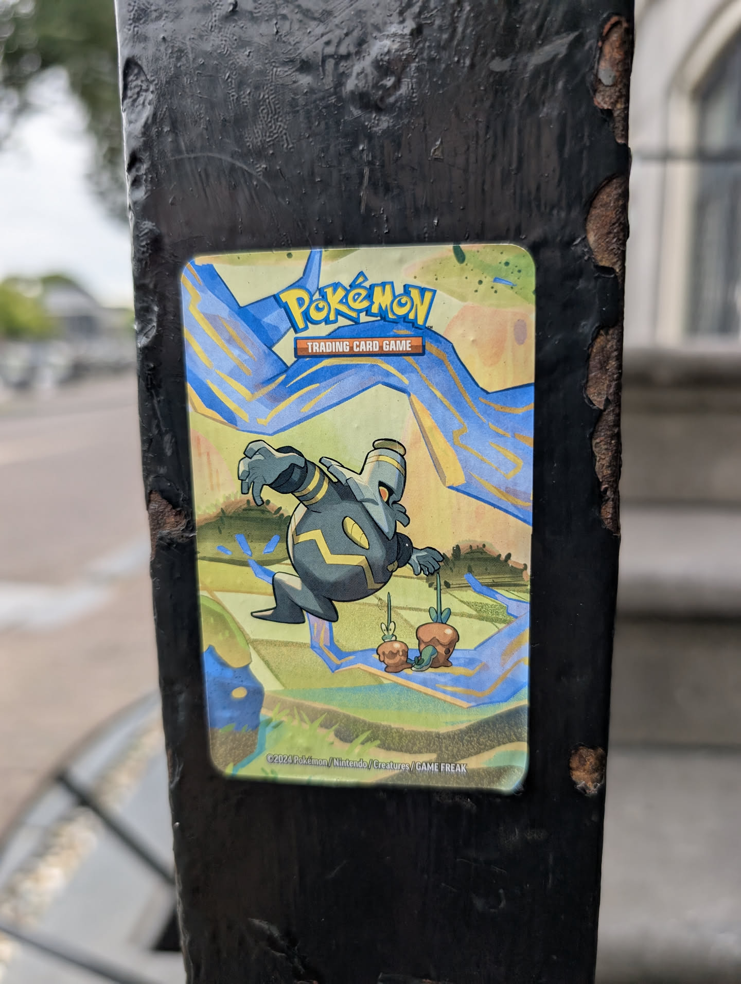 The sticker features the Pokémon Gimmighoul in its chest form, crouched low on a grassy, multi-level landscape. Gimmighoul is predominantly dark gray with yellow accents, and it's holding a small treasure chest in its right hand. The background is a vibrant mix of yellow and blue, with streaks of electric-blue energy radiating outwards. The top of the sticker prominently displays the "Pokémon Trading Card Game" logo. The bottom edge of the sticker includes a copyright notice: "©2024 Pokémon / Nintendo / Creatures / GAME FREAK".