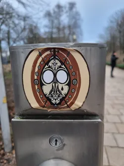 Street sticker A sticker featuring a stylized face with tribal-like patterns. The face is depicted in a circular frame with additional geometric designs. The color scheme is primarily brown and beige. The sticker is slightly peeled from the edges, indicating some wear and tear. It is affixed to a stainless steel pole outside.