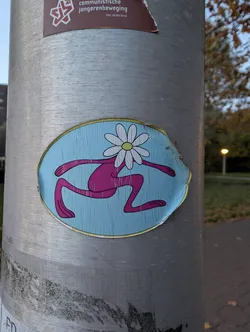 Street sticker The sticker is oval-shaped with a light blue background featuring a subtle wood-grain texture. In the center, a stylized pink figure is depicted in a running or leaping pose. The figure has long, thin limbs and no discernible facial features. Instead, a white daisy with a yellow center serves as its head, with petals extending outwards. The sticker has a yellow border and shows signs of wear and tear around the edges, indicating it has been affixed to the pole for some time.