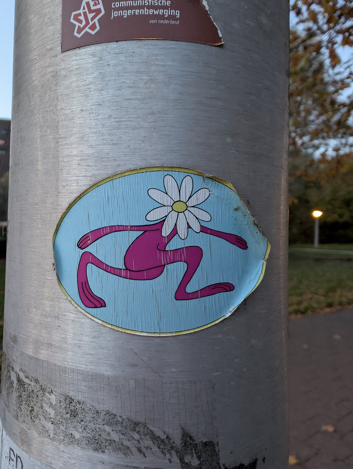 Street sticker The sticker is oval-shaped with a light blue background featuring a subtle wood-grain texture. In the center, a stylized pink figure is depicted in a running or leaping pose. The figure has long, thin limbs and no discernible facial features. Instead, a white daisy with a yellow center serves as its head, with petals extending outwards. The sticker has a yellow border and shows signs of wear and tear around the edges, indicating it has been affixed to the pole for some time.