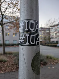 Street sticker The sticker is rectangular with a black background and white text. It features a prominent hashtag symbol '#' followed by the letters 'IUA'. The text is large, bold, and sans-serif. There appear to be two identical stickers stacked vertically, with a thin green border separating them. The bottom sticker has some additional smaller text or symbols below the main text, which are difficult to discern clearly but appear to be '99'. The sticker is applied to a metallic pole.