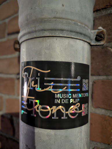 A holographic sticker with the text "Tri-Tone Music Mentors in de Pijp" is affixed to a gray, metallic pipe against a brick wall. The sticker also contains two QR codes. The design is elegant, with a musical note incorporated into the text.