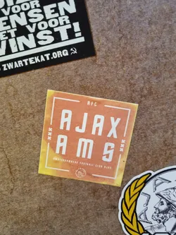 A sticker with a watercolor-like effect depicting the text "AJAX AMS" with a subtle orange and yellow gradient, framed with white lines. The top right corner shows "AFC" in a smaller font, and the bottom has small text in another language. The text translates from Dutch to English as "Amsterdam Football Club Ajax".