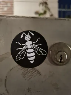 Street sticker A round black and white sticker with an illustration of a bee on a dark background. The bee is depicted in a simple, almost cartoonish style, and is primarily white with some black details. The sticker is affixed to a metallic surface.