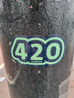 A light green and dark blue sticker with the number 420 in a stylized font. The word 'MASCOTTE' is visible at the bottom. The sticker appears to be affixed to a wet, dark-colored pole.