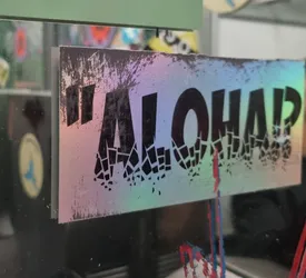 Street sticker This eye-catching sticker features the word "ALOHA!" in black lettering, set against a holographic rainbow background. The letters appear to be breaking apart into smaller pieces, adding a dynamic visual effect.  The sticker has a slightly worn or distressed look around the edges. 