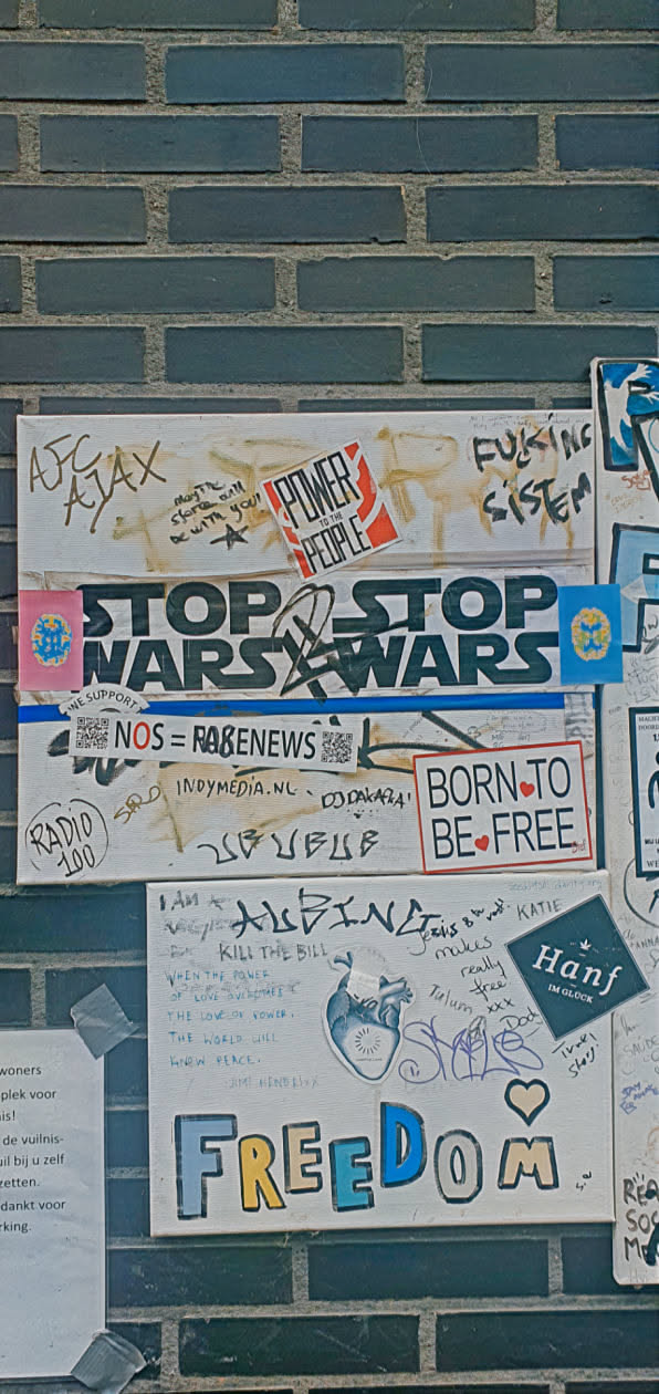 The image shows a collection of stickers and writings on what appears to be a brick wall. A prominent sticker reads "STOP WARS", while another states "POWER TO THE PEOPLE". There is also a sticker with the text "BORN TO BE FREE". Other writings include "FREEDOM", "KILL THE BILL", and various signatures and messages.