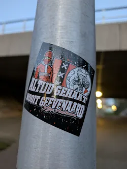 Street sticker The sticker is predominantly black with red and white accents. On the left, it features a stylized illustration of a person wearing a red balaclava and a red jacket with white trim. To the right of this figure, there is a representation of the Ajax Amsterdam crest, which includes three white crosses and a depiction of Ajax himself. Below these images, bold white text reads 'ALTIJD GEHAAT' and 'NOOIT GEEVEE NAARD', with 'Ajax Amsterdam' written in a stylized script below. The background of the sticker has a distressed, splattered effect in red and white.
