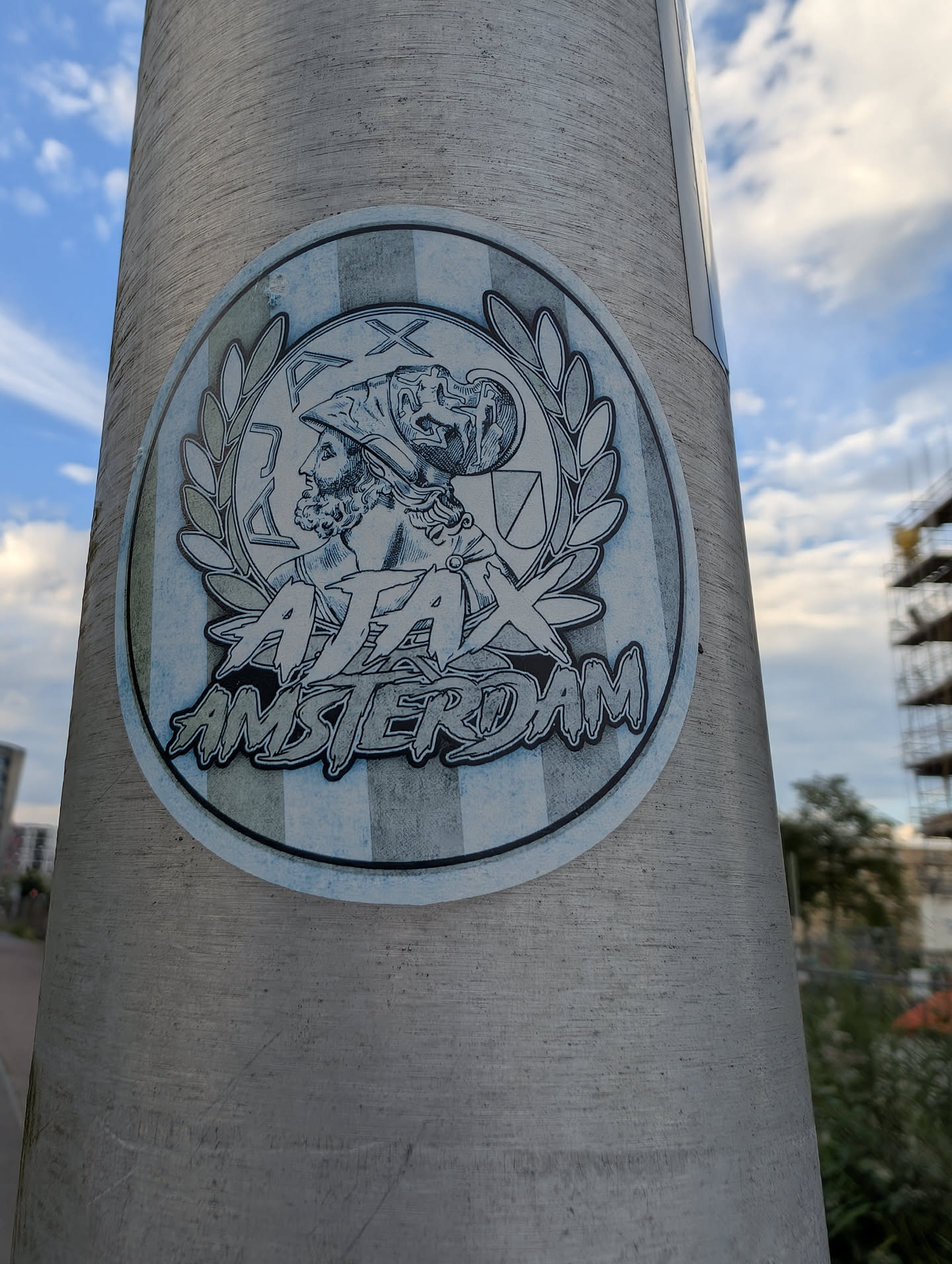 An oval sticker featuring a blue-toned illustration of the Greek hero Ajax in profile with a helmet, framed by a laurel wreath. The text 'AJAX AMSTERDAM' is written below in a stylized graffiti font against a striped background.