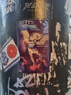 Street sticker This sticker shows a child with angel wings holding a flag, triumphing over a fallen adversary.  The style is reminiscent of vintage or classical artwork. The number 12 is visible on the child's clothing, and a website address, www.manchesterglory.com, is visible in the lower portion of the sticker. 