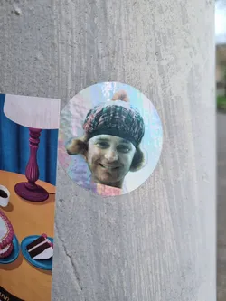 A round holographic sticker featuring a person wearing a tam o' shanter. The person has shoulder-length hair and a happy expression. The background of the sticker seems to have a rainbow effect.