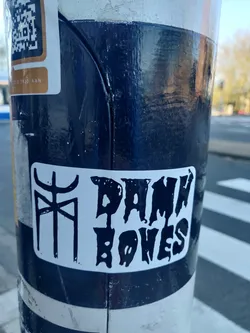 Street sticker A black and white sticker with the text "Damn Bones" in a stylized font. There is also a smaller symbol to the left of the text. The sticker is affixed to a dark blue pole.