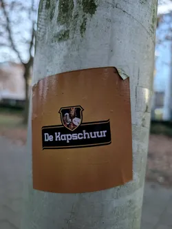 Street sticker The sticker is predominantly a warm, gradient brown, transitioning from a lighter shade at the top to a darker shade at the bottom. Centered on the sticker is a black shield-shaped emblem. Within this emblem, a small, detailed illustration depicts a king wearing a crown and holding a frothy beer mug. Below the emblem, a black banner with a thin gold outline contains the white text 'De Kapschuur'. The sticker appears to be slightly peeling at the top right corner.