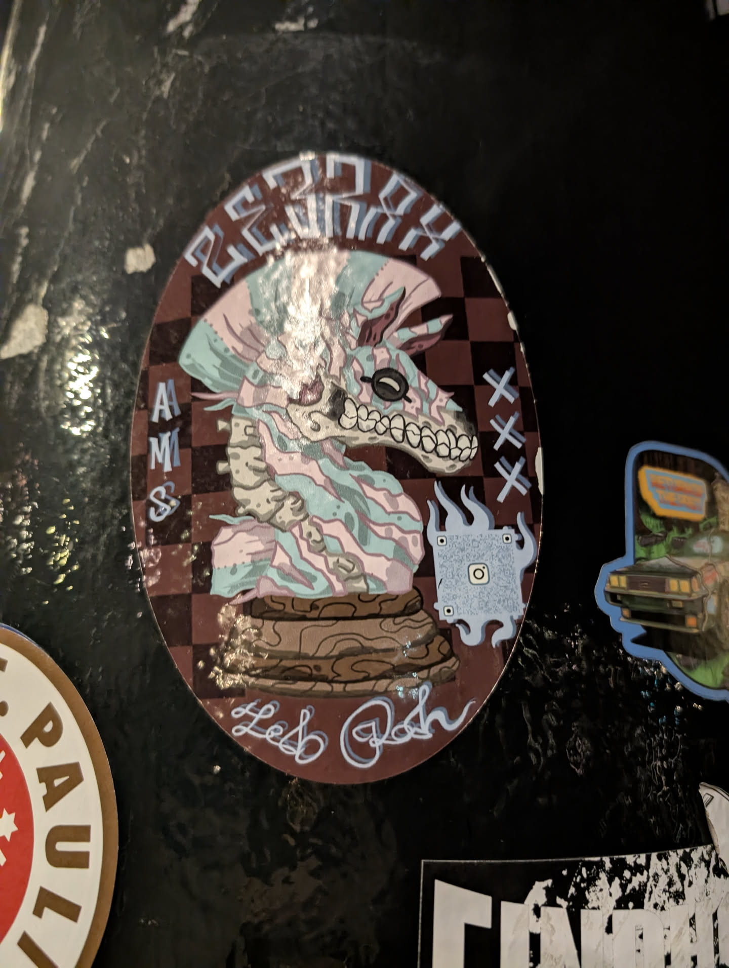 Street sticker The central sticker is oval-shaped, featuring a highly stylized, skeletal horse head reminiscent of a chess knight piece. The creature is rendered in a striking palette of light pink, mint green, and grey, with black outlines, giving it an ethereal and slightly macabre appearance. Its eye sockets are empty, but it wears small, round, dark-rimmed glasses, and its jaw reveals a row of sharp teeth. The horse's mane and body are depicted with flowing, wavy patterns. The background is a dark brown and black checkered pattern, evoking a chessboard. The horse rests on a textured, wood-like base. Text elements include "BEROXX" in stylized block letters at the top, "AMSS" vertically on the left, and "Les Rosh" in a flowing script at the bottom. On the right side, there are three 'X' marks. A light blue, flame-like or smoke-like graphic on the bottom right corner contains a QR code with a distinct Instagram logo embedded in its center. The overall aesthetic blends fantasy, street art, and a touch of the macabre.