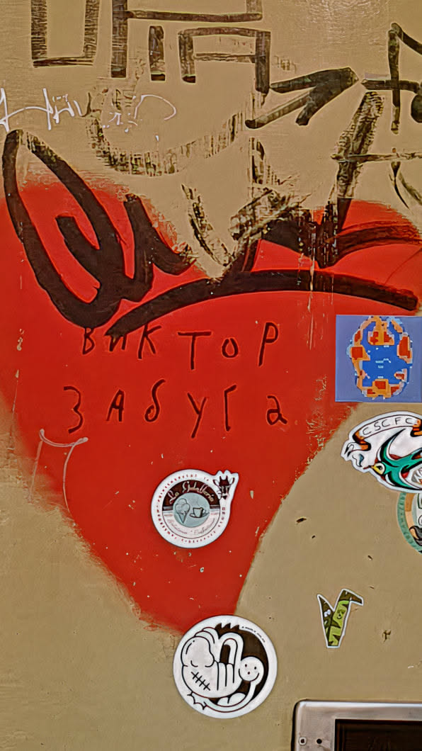 A view of an urban wall featuring several stickers. The most prominent is a red, heart-shaped spray painting with the text "Виктор Забуга" written in black on the heart. Above the heart, there are some dark abstract strokes. There's a round sticker with an image of a coffee and ice cream cup with the text "... La Gelateria...". Another one depicting a smiling baby-like face is also there. In addition, there is a pixel art sticker. Other graffiti and markings contribute to the gritty, urban feel.
