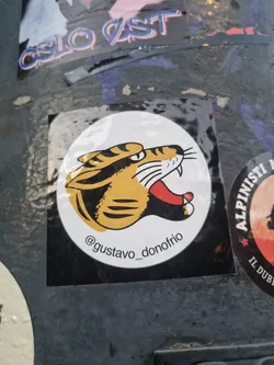 A round sticker featuring a vintage-style illustration of a tiger's head in yellow and black. The background is black and the artist's Instagram handle is visible at the bottom.