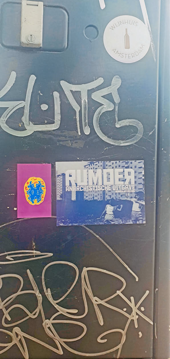The Rumoer sticker has a blue background and white text that reads "RUMoer ANARCHISTISCHE UITGAVE rumoer.noblogs.org". The image on the sticker shows people in front of a building. The metal surface also has other stickers, including one with a brain graphic and one with a wine bottle logo, as well as white graffiti.