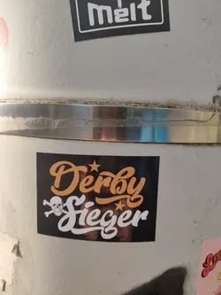 Street sticker A black sticker with the text "Derby Sieger" in orange and white stylized lettering.  A skull and crossbones is featured below the text. The sticker appears to be affixed to a metal surface.