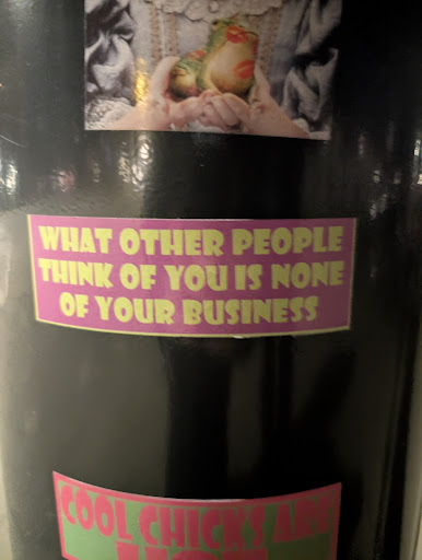 A rectangular sticker with a pink background and yellow text saying "WHAT OTHER PEOPLE THINK OF YOU IS NONE OF YOUR BUSINESS". It is stuck on a dark surface.