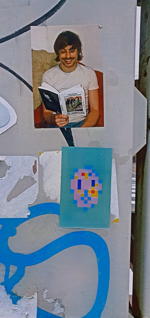 A turquoise sticker featuring a pixelated face, made up of pink, blue, yellow, and red squares, is adhered to a weathered wall.
