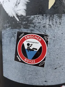Street sticker A round sticker with a red border and a black center. It has a pictogram of a person falling into water and the text EMSCHER Lebensgefahr! (EMSCHER Danger of death!). There is also the text #ABAE BOYZZ at the bottom. The sticker is affixed to a dark-colored surface.